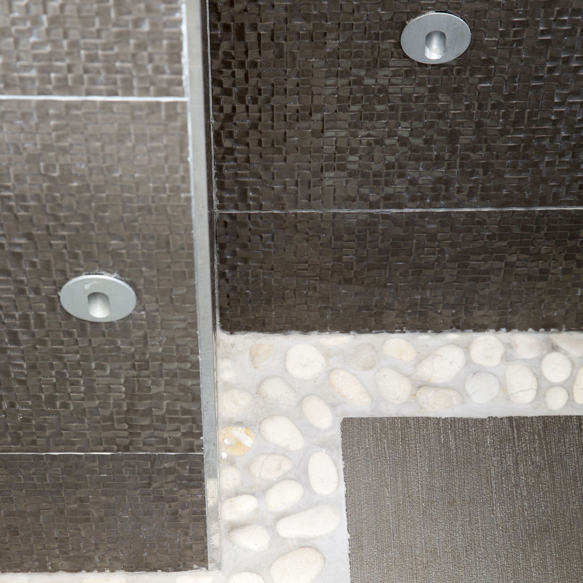 iluminacion en duchas: primer plano de azulejos de baño metálicos marrones, suelo de efecto guijarro, foco de pared